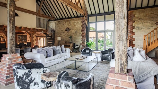 A spacious living area with a high ceiling, exposed wooden beams, and brick walls. There is a white sofa, a glass coffee table, and several leather armchairs. A staircase with a wooden railing is visible in the background. - Stunning 6 bedroom converted barn nestled in Souths Downs; 5,000+ sq ft; 9 acres (Storrington)