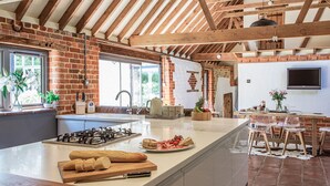 A kitchen with a brick wall, wooden beams, a large island, and a dining area with chairs. - Stunning 6 bedroom converted barn nestled in Souths Downs; 5,000+ sq ft; 9 acres (Storrington)