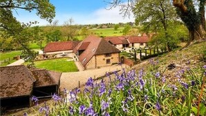 A rural setting with traditional stone buildings, a grassy field, and blooming purple flowers. - Stunning 6 bedroom converted barn nestled in Souths Downs; 5,000+ sq ft; 9 acres (Storrington)