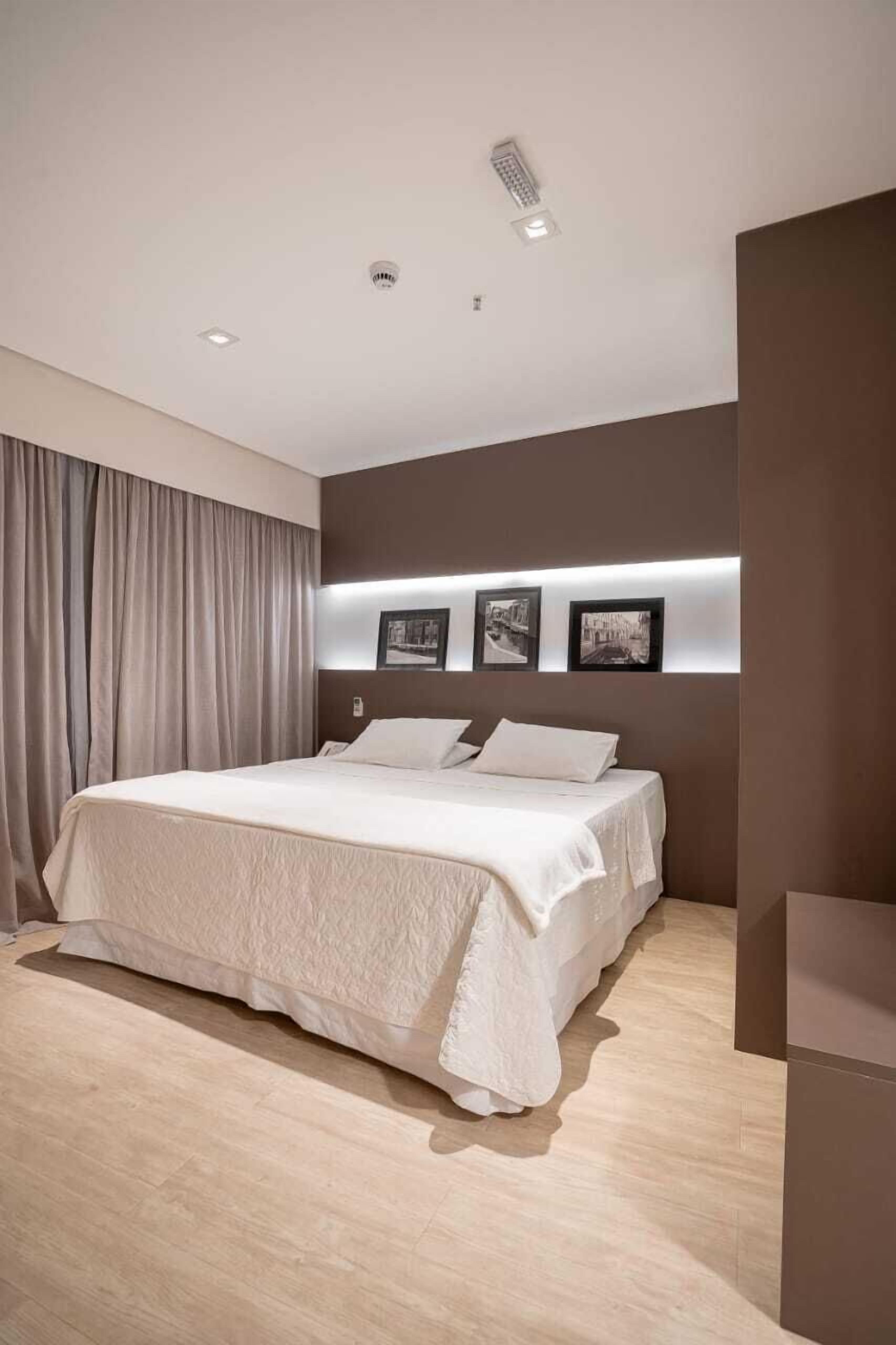 Une chambre à coucher moderne avec un grand lit, une tête de lit dotée de lampes fixées au mur et un rideau sur le côté gauche.