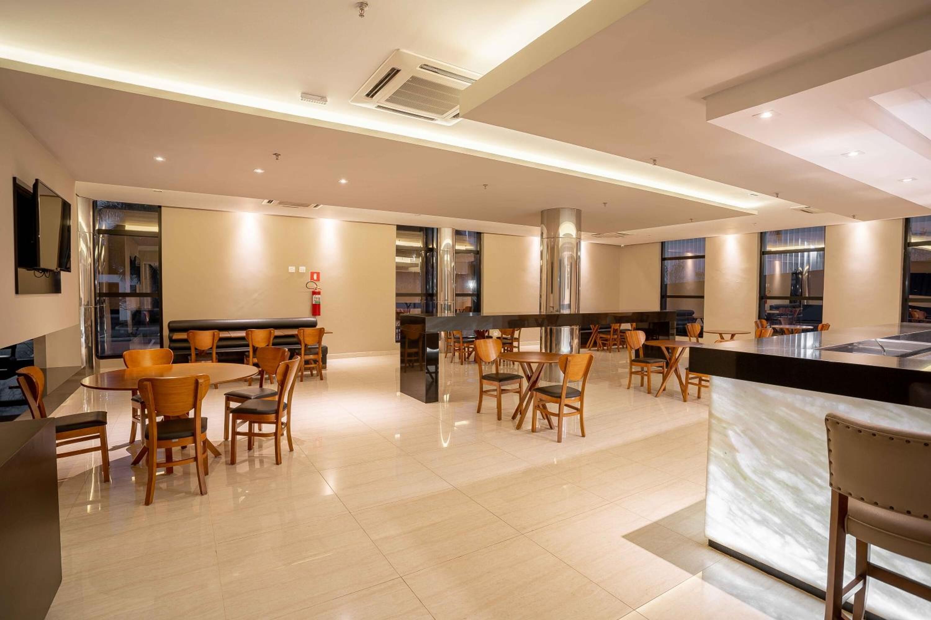 Una hall d'hotel moderna con un'area bar centrale, diversi tipi di posti a sedere e una grande cucina sullo sfondo.