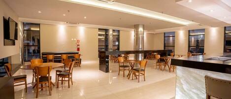 Una hall d'hotel moderna con un'area bar centrale, diversi tipi di posti a sedere e una grande cucina sullo sfondo.