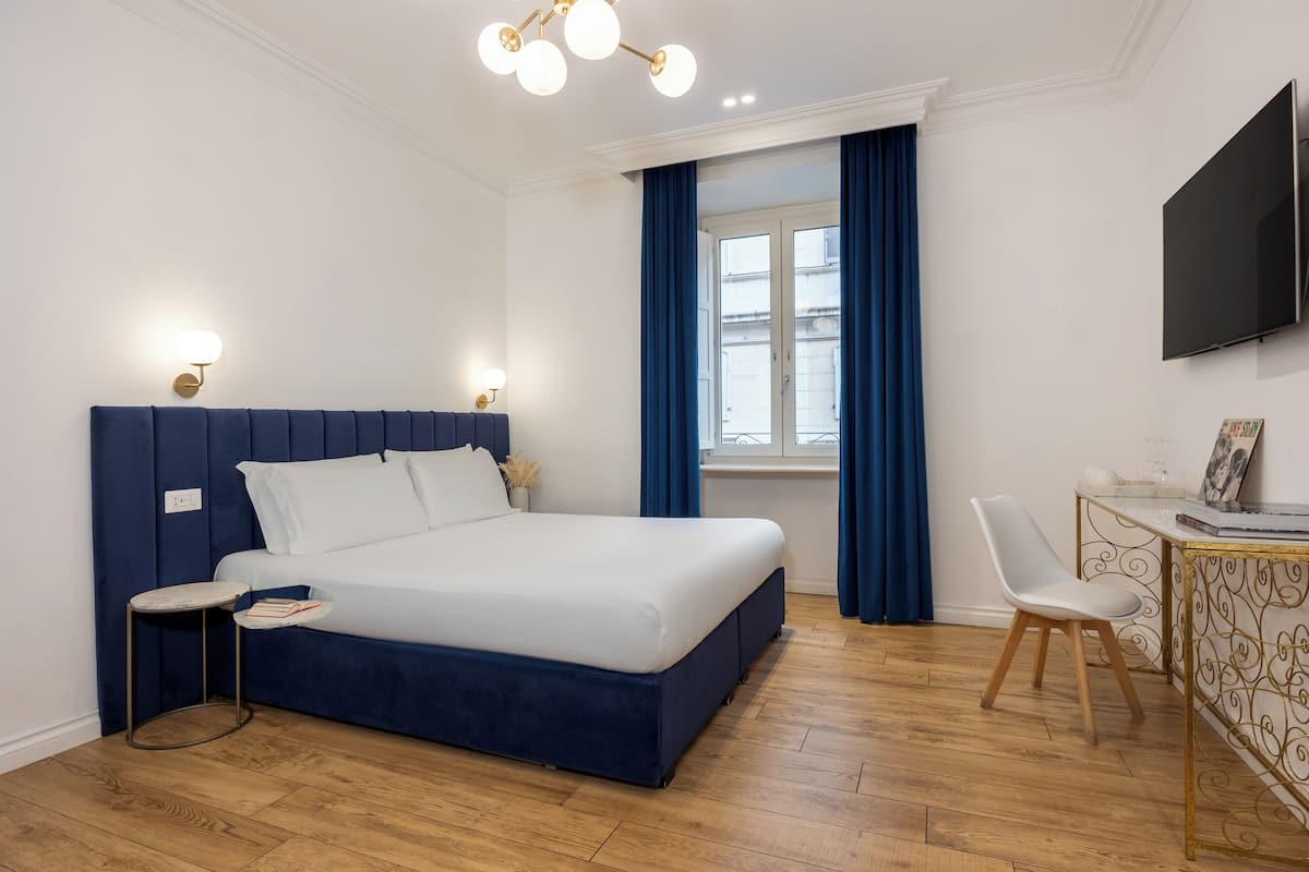 A modern bedroom with a large bed, bedside tables, a desk with a chair, a TV, and a window with curtains.