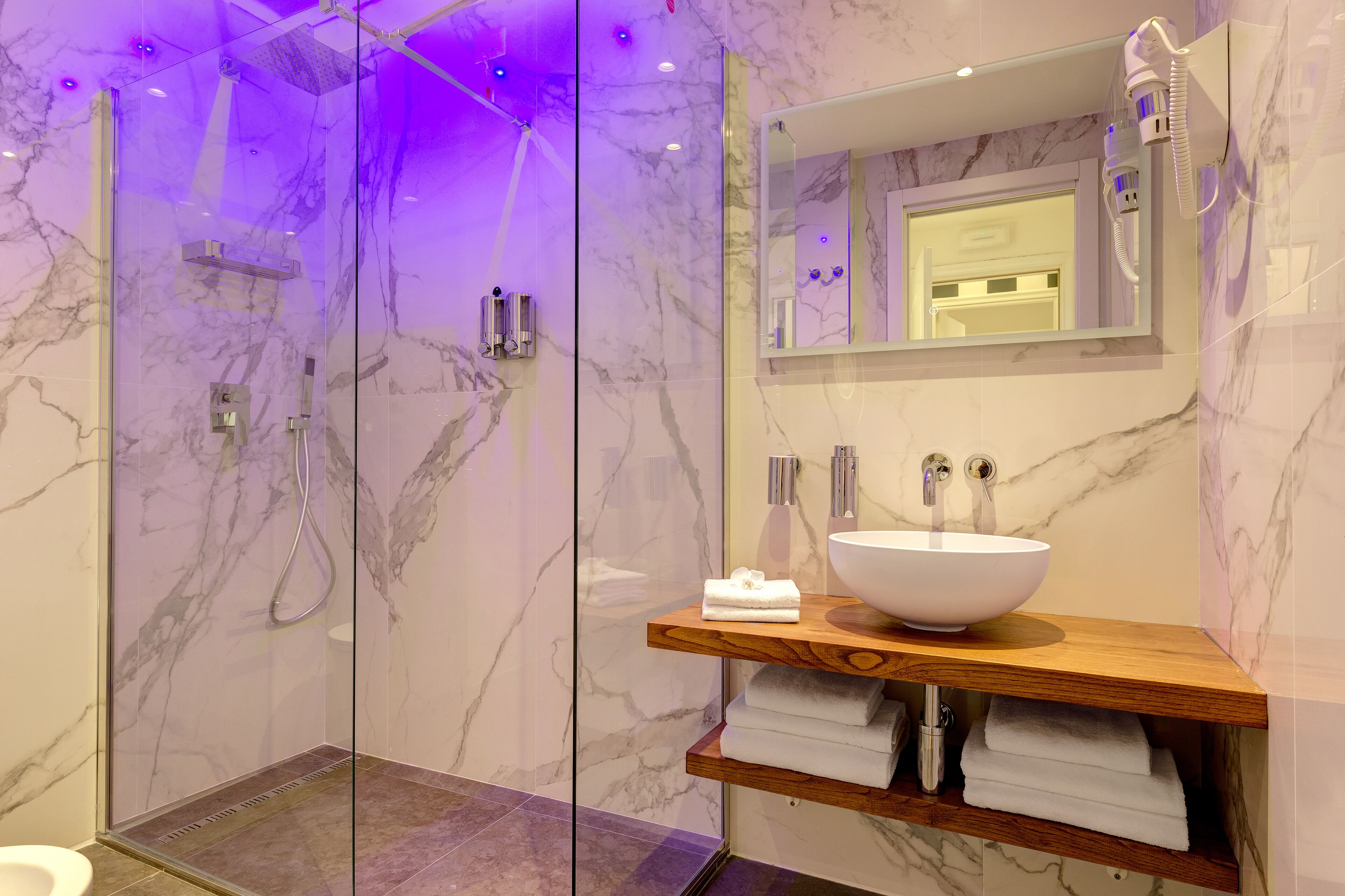 A modern bathroom with a glass-enclosed shower, a marble-tiled wall, a wooden vanity with a large sink, and a mirror above the vanity.