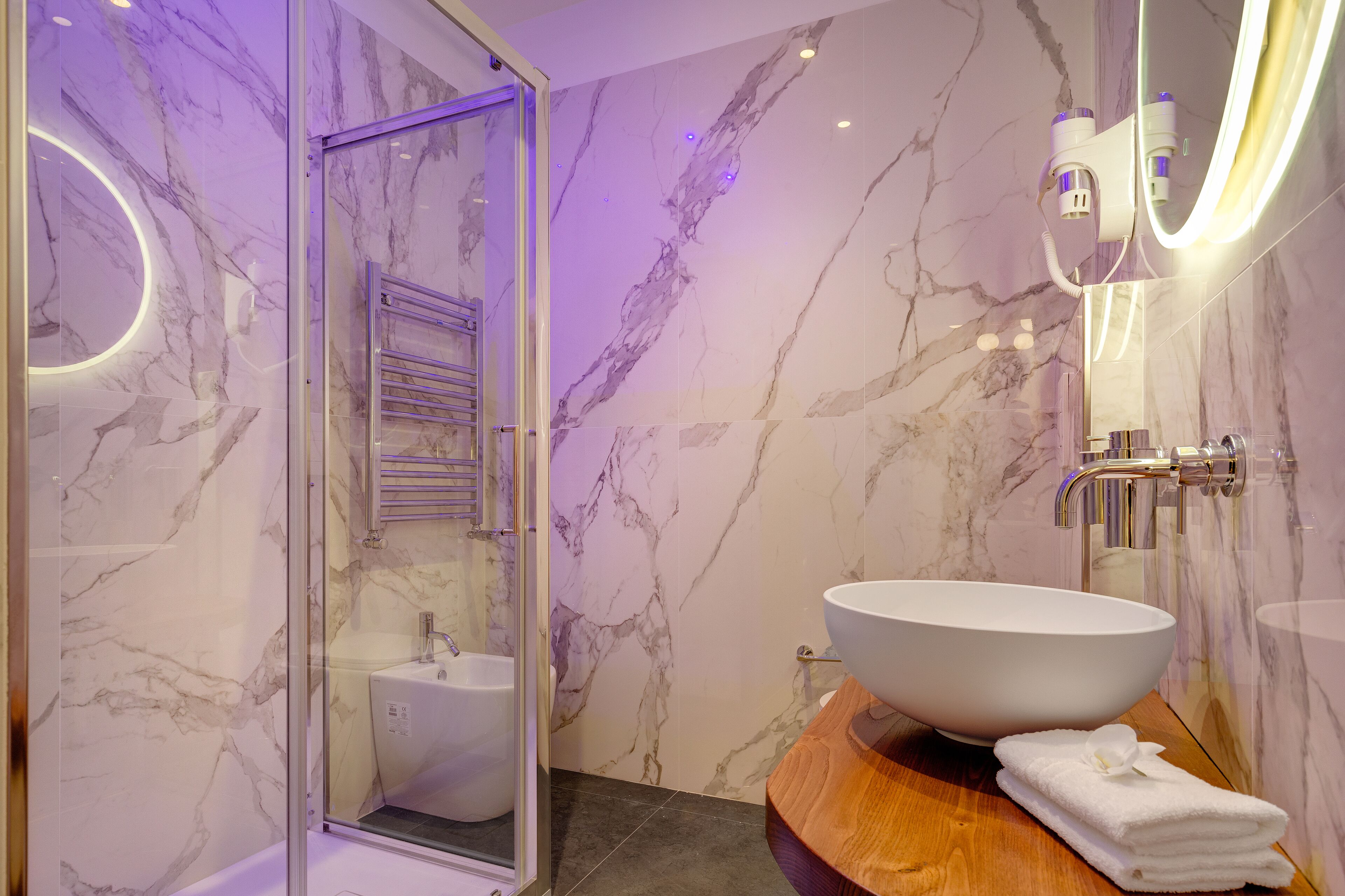 A modern bathroom with a glass shower enclosure, a marble backsplash, a large white vessel sink, and a wooden vanity.