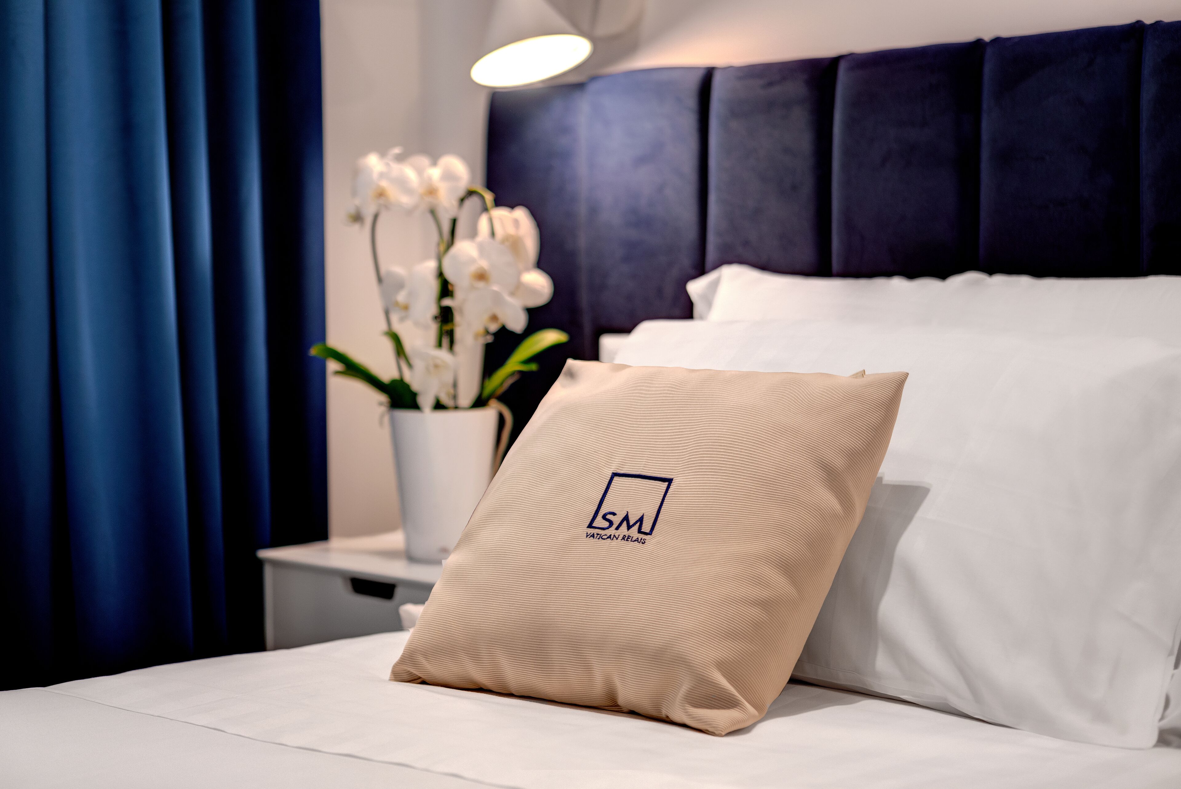 A neatly made bed with a pillow featuring a logo, a vase with white flowers, and a blue curtain.