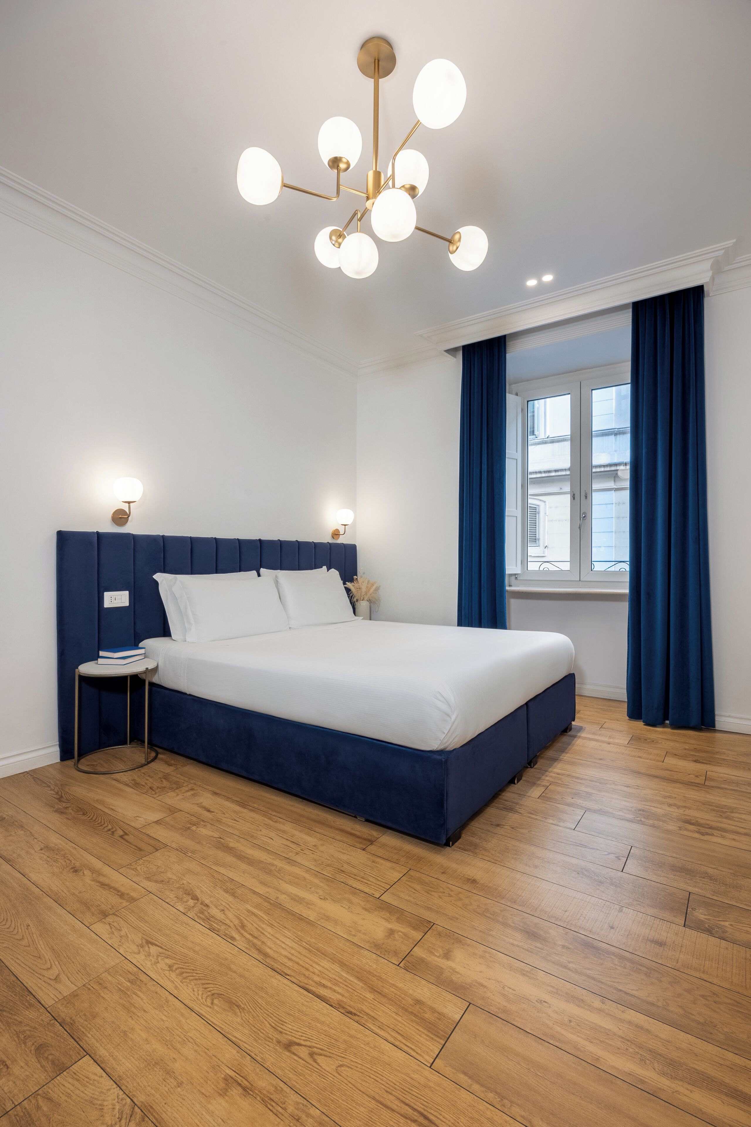 A bedroom with a large bed, a nightstand, a modern chandelier, and a window with curtains.
