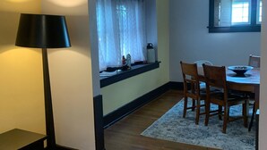 A dining area with a table and chairs, a window with curtains, a lamp, and a small shelf with items.