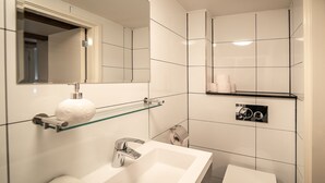 A modern bathroom with a white sink, mirror, and toilet. There is a glass shelf above the sink holding a roll of toilet paper and a white bath mat. - Dutch House (Amsterdam)