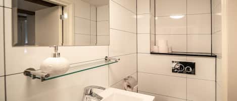 A modern bathroom with a white sink, mirror, and toilet. There is a glass shelf above the sink holding a roll of toilet paper and a white bath mat.