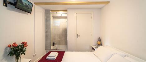 A hotel room with a bed, a small table with a vase of flowers, a shower area, and a television mounted on the wall.
