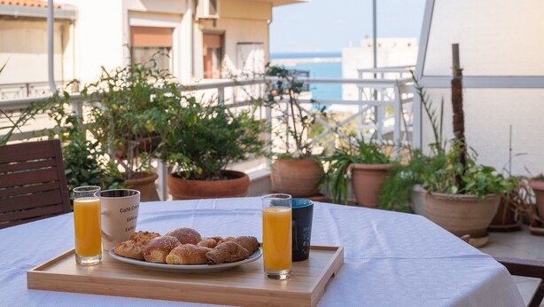 Una mesa con una bandeja de cruasanes, dos vasos de jugo de naranja y una taza de café en un balcón con vistas a la ciudad y al mar.