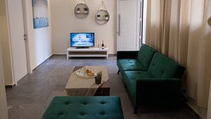 Una sala de estar con un sofá verde, un televisor sobre un mueble blanco, una mesita con una botella y vasos, y una puerta con marco blanco.