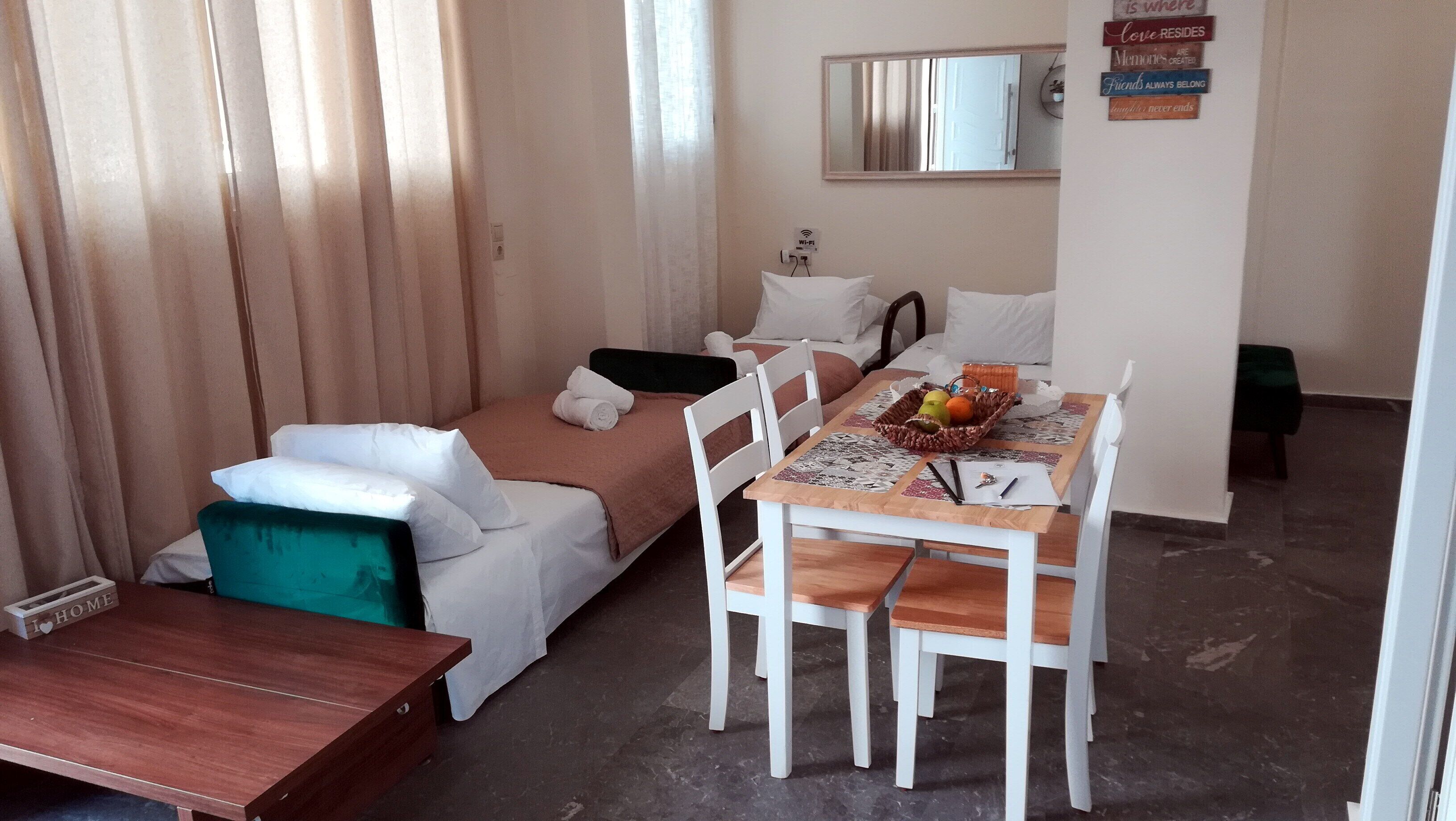 A small, well-lit room with a bed, a wooden table with chairs, and a dining table set with fruits and a basket.