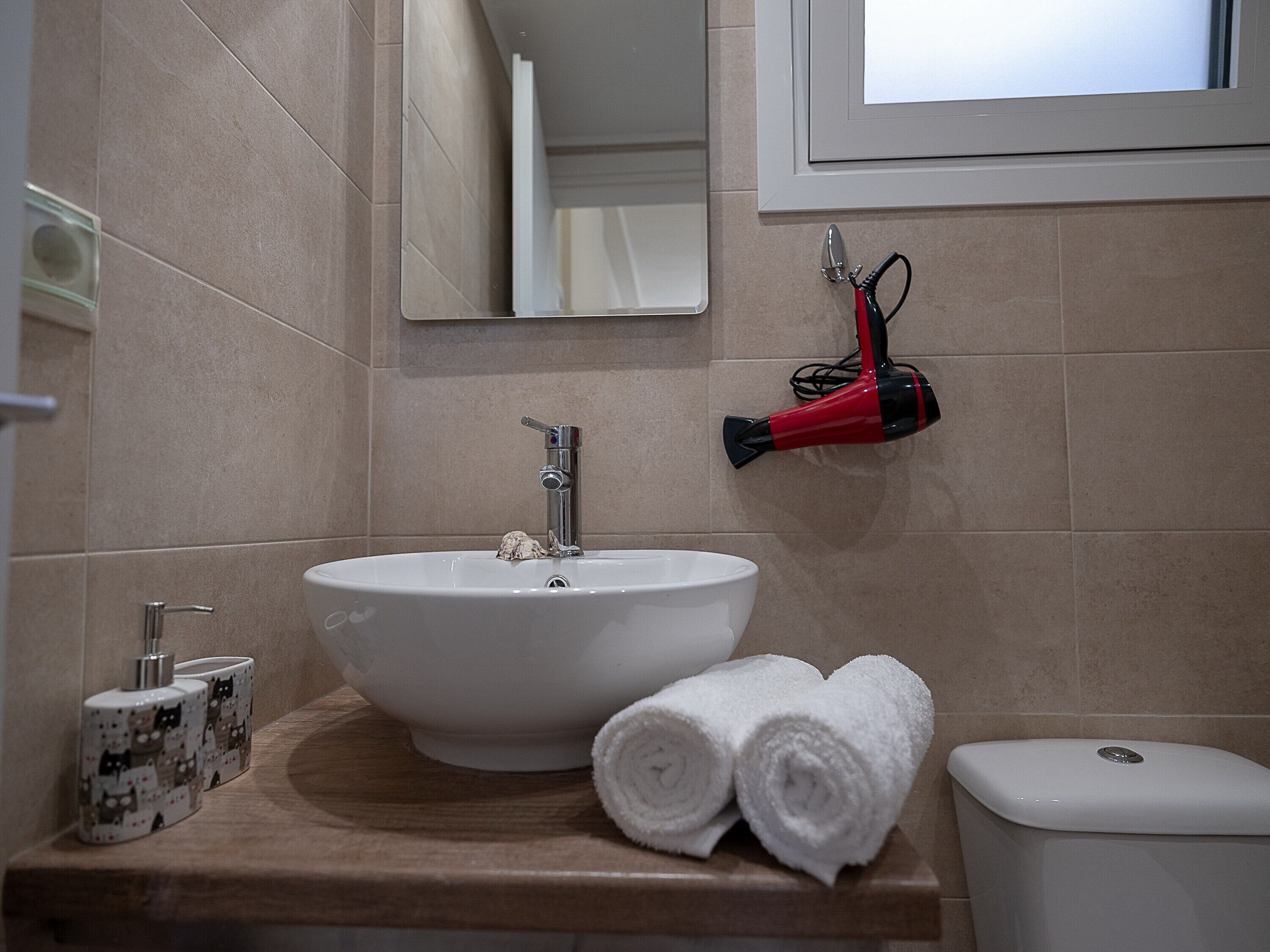 Un baño con un lavabo blanco, un espejo, un secador de pelo rojo y dos toallas enrolladas sobre una encimera de madera.
