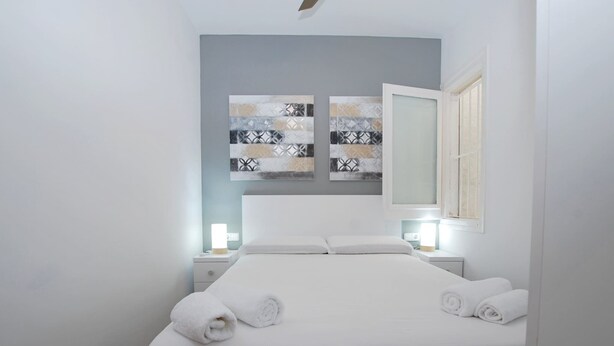 Una cama bien tendida con sábanas blancas y dos toallas enrolladas, una mesita de noche con una lámpara, y un espejo grande en la pared.