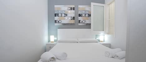 A neatly made bed with white linens and two rolled towels, a bedside table with a lamp, and a large mirror on the wall.
