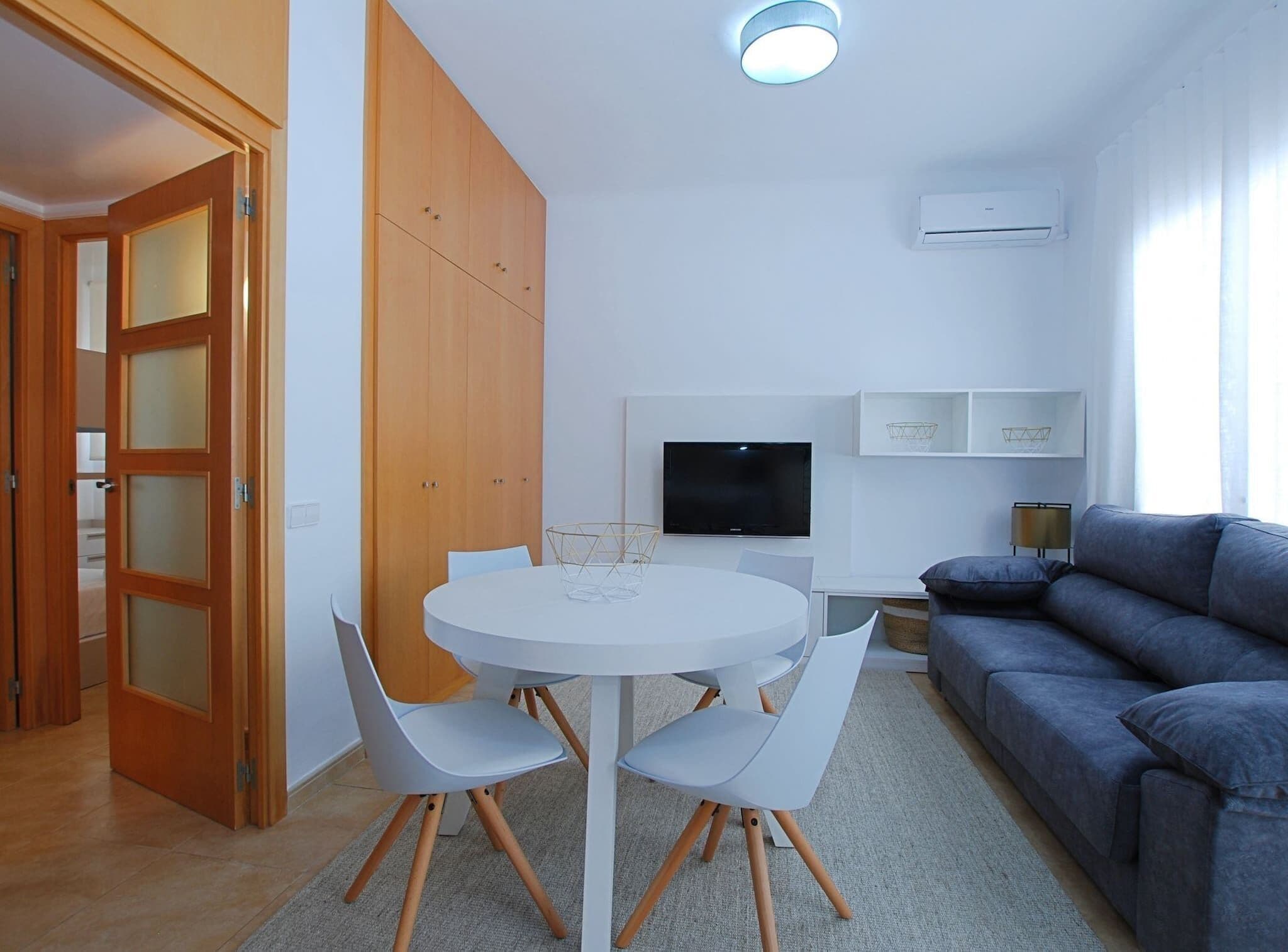 Una sala moderna con una mesa redonda blanca, dos sillas blancas, un sofá gris, un televisor de pantalla plana y una lámpara de techo.