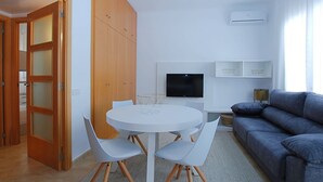 Una sala moderna con una mesa redonda blanca, dos sillas blancas, un sofá gris, un televisor de pantalla plana y una lámpara de techo.