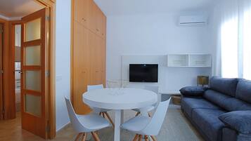 Una sala moderna con una mesa redonda blanca, dos sillas blancas, un sofá gris, un televisor de pantalla plana y una lámpara de techo.