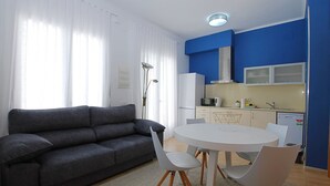 Una sala moderna con un sofá gris, una mesa de comedor redonda con sillas blancas y una cocina con paredes azules y armarios blancos.