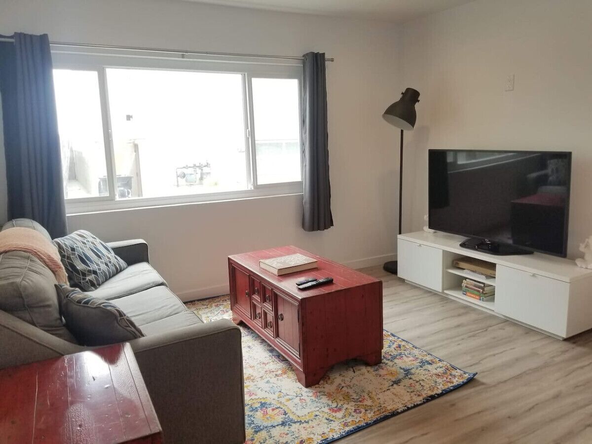 Een woonkamer met een bank, een rode salontafel, een televisie op een witte dressoir en een raam met gordijnen.