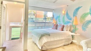 A bedroom with a bed, a ceiling fan, a window with a view of buildings, and a wall with leaf patterns. - El Sol Beachfront + Pool + Beach + Parking (San Juan)