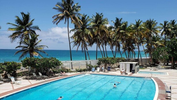 A swimming pool with clear blue water, surrounded by palm trees and a view of the ocean. - El Sol Beachfront + Pool + Beach + Parking (San Juan)