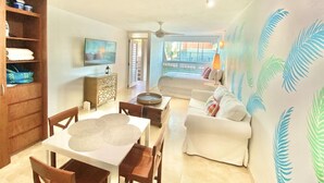A spacious living room with a sofa, dining table, and chairs, a television, and a large window with a view. - El Sol Beachfront + Pool + Beach + Parking (San Juan)