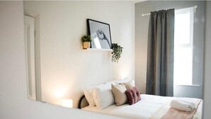 Un lit bien fait, avec des oreillers et une couverture, une petite étagère avec une plante, un tableau encadré au mur et une fenêtre avec des rideaux.