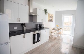 A modern kitchen with white cabinets, a black countertop, and a dining area with a table and chairs. - Buchanan House Sasco Apartments (Blackpool)