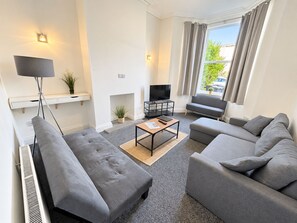 A modern living room with a sofa set, a flat-screen TV, a fireplace, and a large window with curtains. - Buchanan House Sasco Apartments (Blackpool)