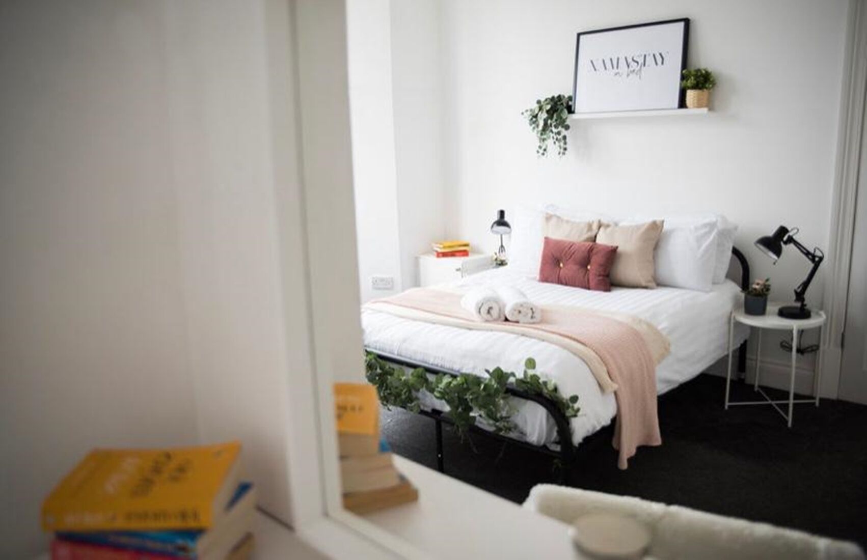 A bedroom with a bed, bedside tables, a wall-mounted shelf with a plant, and a framed sign above the bed.