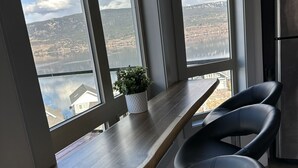 Moderni keittiö, jossa on puinen työtaso, kaksi mustaa tuolia, ruostumattomasta teräksestä valmistettu jääkaappi ja suuret ikkunat, joista avautuu näkymä järvelle ja vuorille.