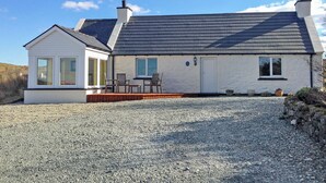 A two-story house with a gravel driveway, a small porch with chairs, and a white exterior. - Teeny’s Cottage is a spacious family holiday home set all on the ground floor. (Portree, near Dunvegan)