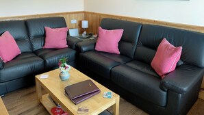 A corner seating area with black leather sofas, pink cushions, a wooden coffee table, a laptop, a vase with flowers, and a painting on the wall. - Teeny’s Cottage is a spacious family holiday home set all on the ground floor. (Portree, near Dunvegan)