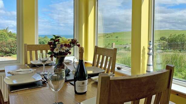 A dining table with chairs, a vase of flowers, a bottle of wine, and a view of green fields and hills through large windows. - Teeny’s Cottage is a spacious family holiday home set all on the ground floor. (Portree, near Dunvegan)