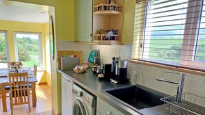 A kitchen with a washing machine, a coffee maker, and a window with blinds. - Teeny’s Cottage is a spacious family holiday home set all on the ground floor. (Portree, near Dunvegan)