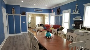 Comedor con mesa de madera y sillas blancas, cocina con armarios blancos y paredes azules decoradas con motivos náuticos.