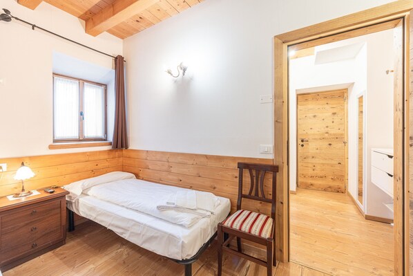 A bedroom with a wooden headboard, a single bed, a nightstand with a lamp, a chair with a striped cushion, and a wooden door leading to another room.