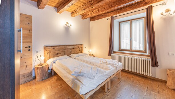 A bedroom with a wooden bed, a wooden floor, a window with curtains, a radiator, and a wooden door. - Alla Regia (Vodo Cadore)