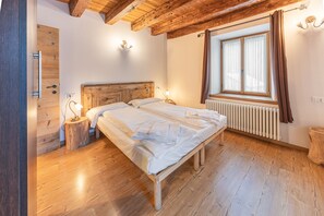 A bedroom with a wooden bed, a wooden floor, a window with curtains, a radiator, and a wooden door.