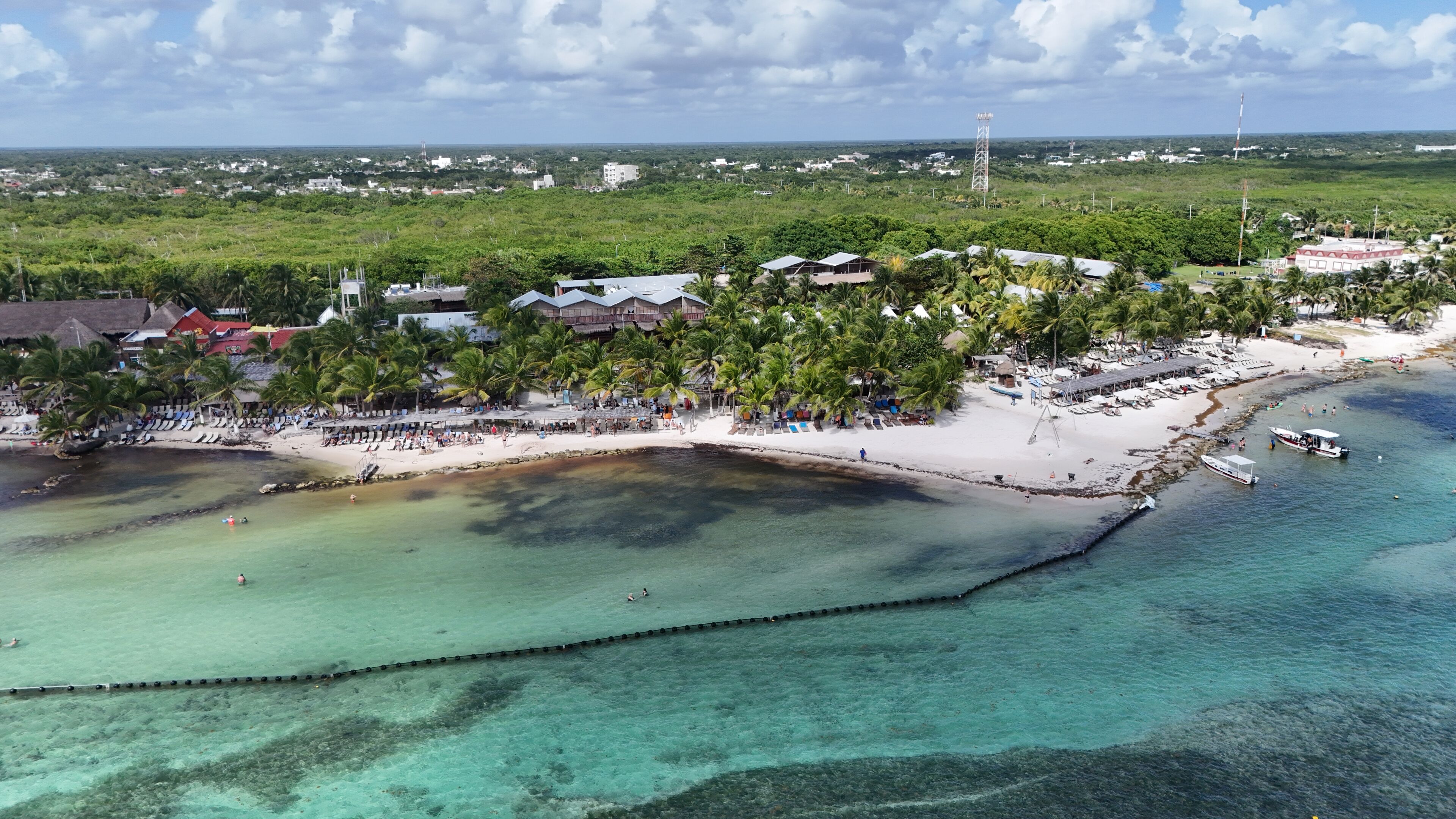 Vista aérea de un resort costero con zonas de playa, palmeras y aguas turquesas cristalinas.