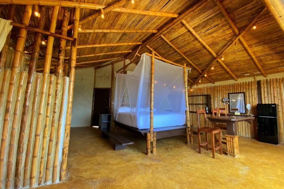 A room with a bed under a mosquito net, bamboo walls, a desk with a chair, and a refrigerator.