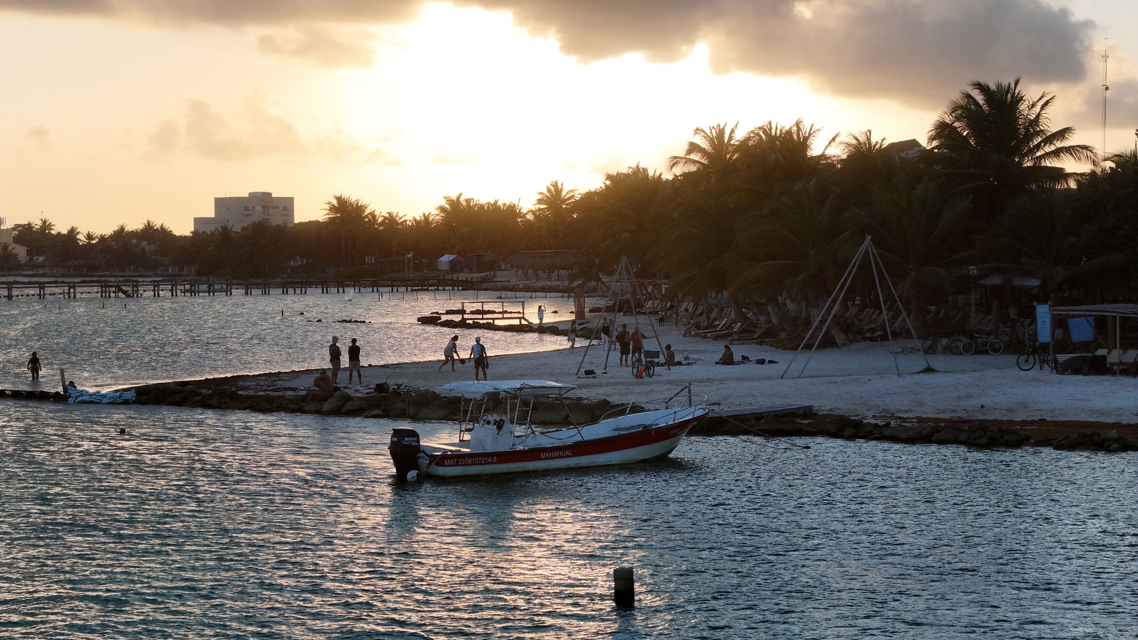 A boat docked at a pier with people on the beach, palm trees, and a sunset.