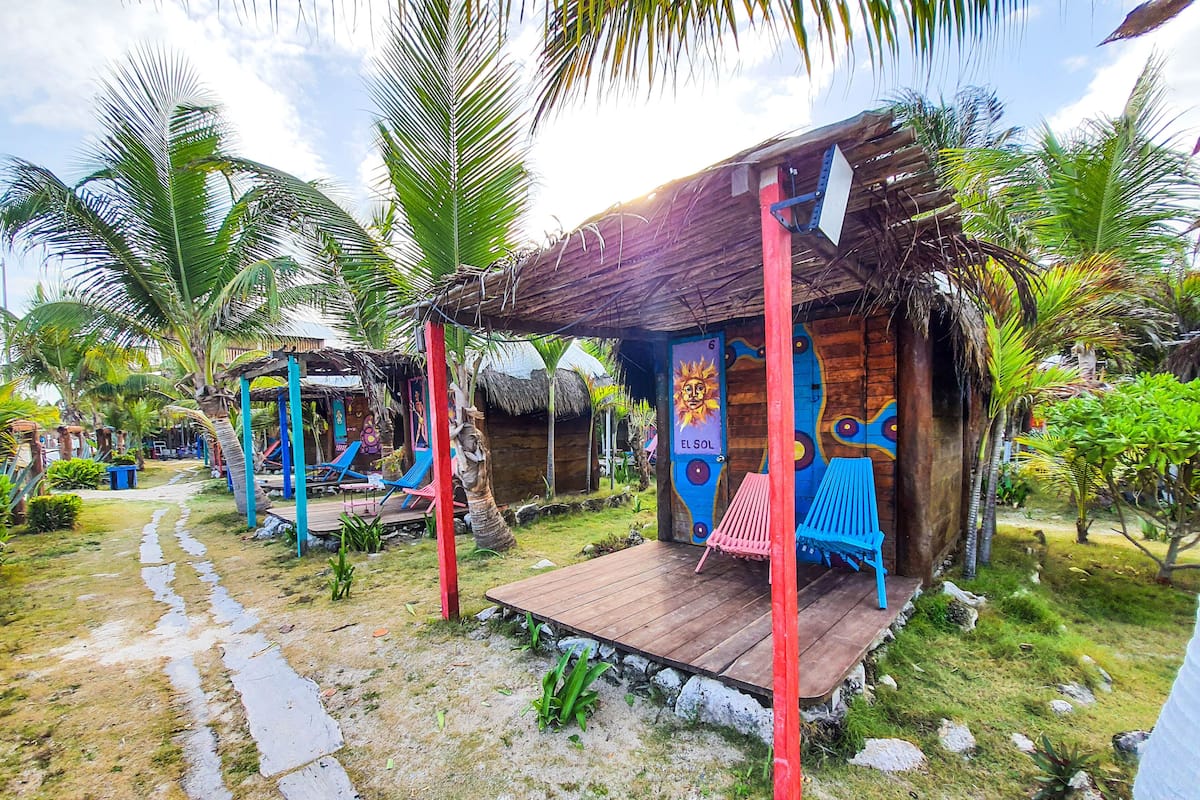 A rustic wooden structure with a thatched roof, a colorful mural, and a small deck area with a hammock.