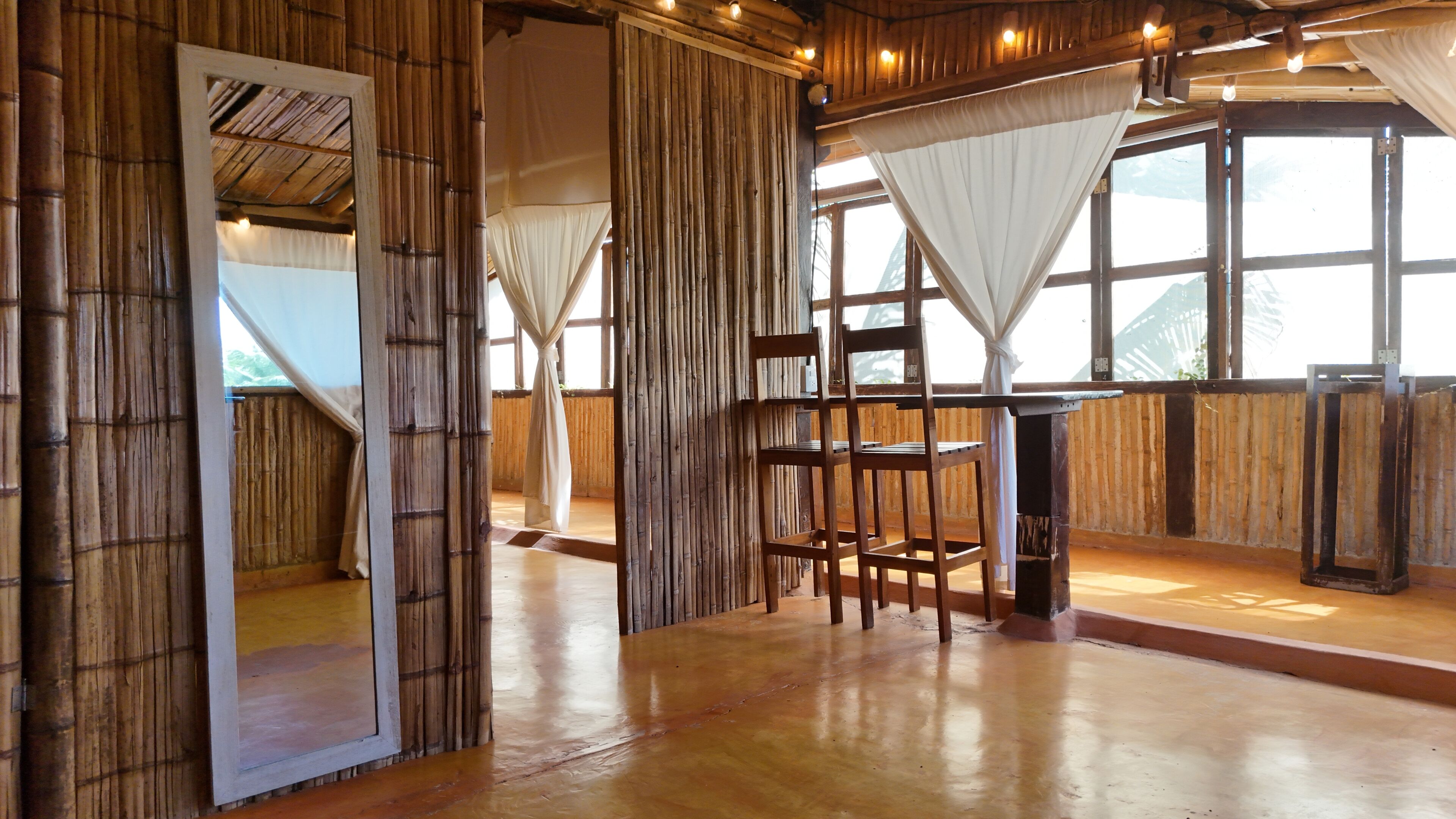 A room with bamboo walls, a wooden chair, a table, and large windows with white curtains.