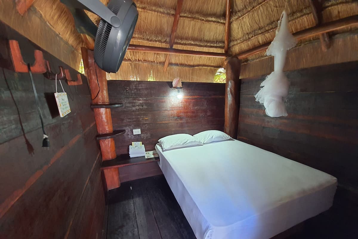 A small, rustic room with a single bed, a wooden wall, a thatched roof, and a wall-mounted fan.