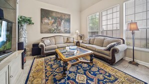 A living room with a sofa, a glass coffee table, a painting on the wall, and a large window with blinds. - Ideally Located 3-Bedroom Townhouse (Tulsa)