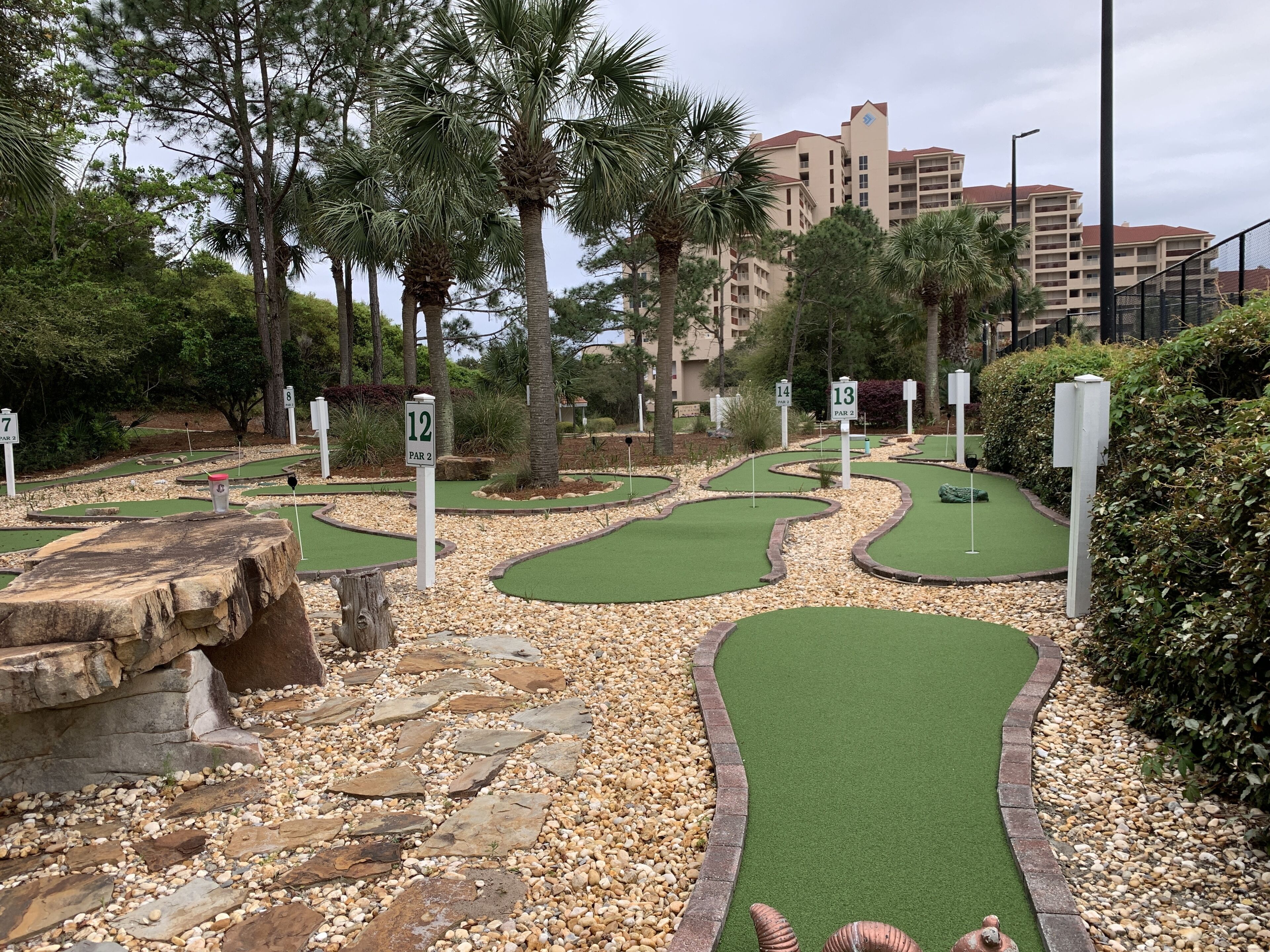 A miniature golf course with numbered holes, surrounded by trees and a building in the background.
